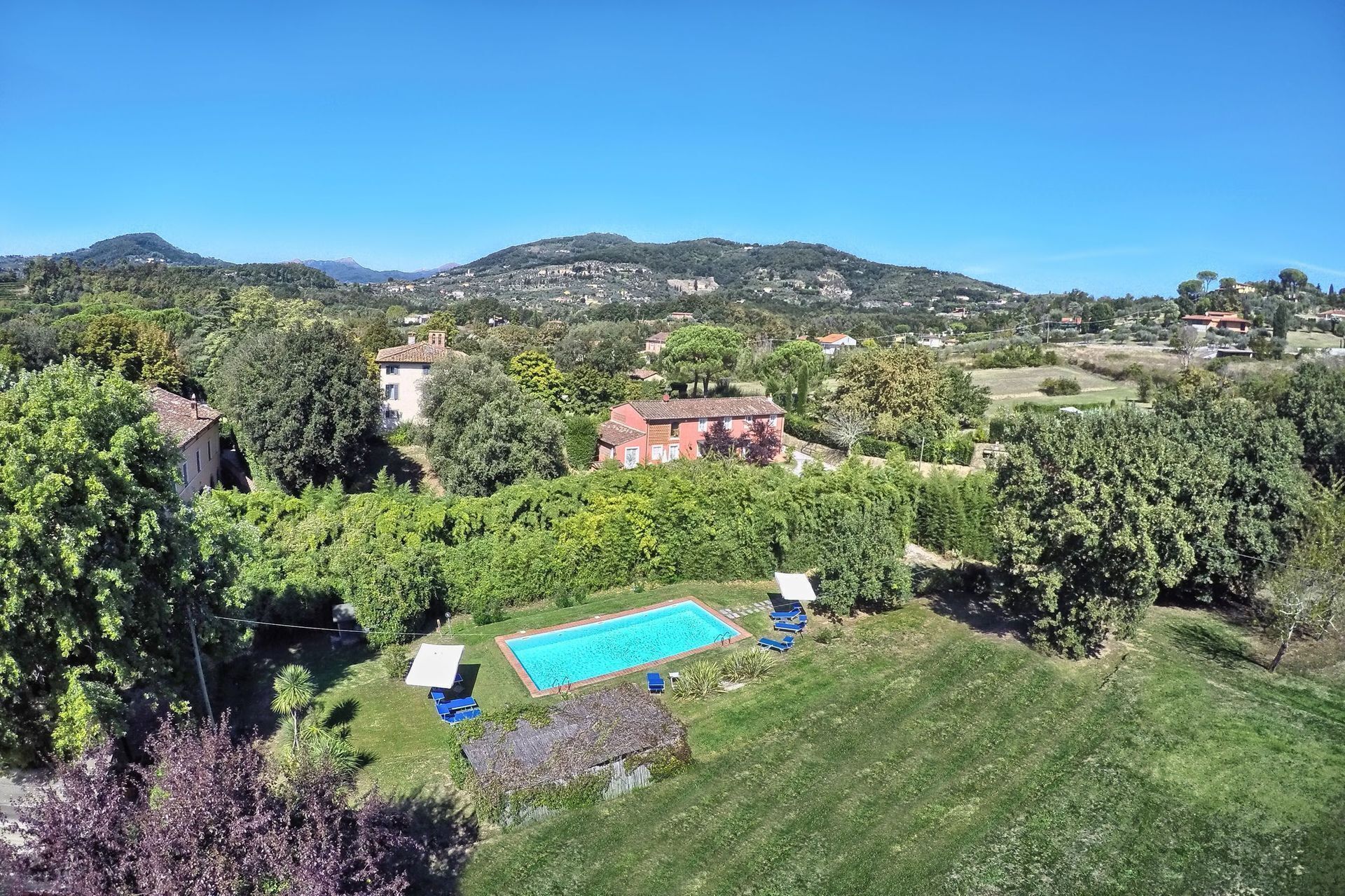 A residential area with a swimming pool surrounded by greenery, houses, and hills in the background.
