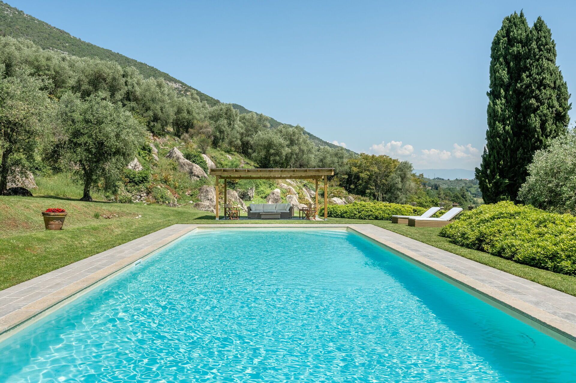 Piscina retangular com pérgola e área de estar ao fundo, rodeada por vegetação exuberante e colinas.
