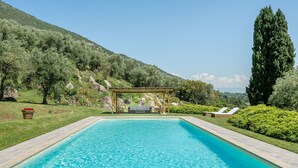 Une piscine rectangulaire, agrémentée d’une pergola et d’un espace détente à l’arrière-plan, le tout entouré d’une végétation luxuriante et de collines.