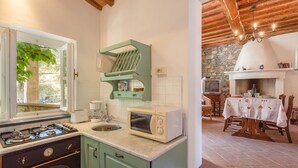 Una cocina con estufa, microondas y un colgador verde. Se aprecia una pared de piedra y una zona de comedor con mesa y sillas.