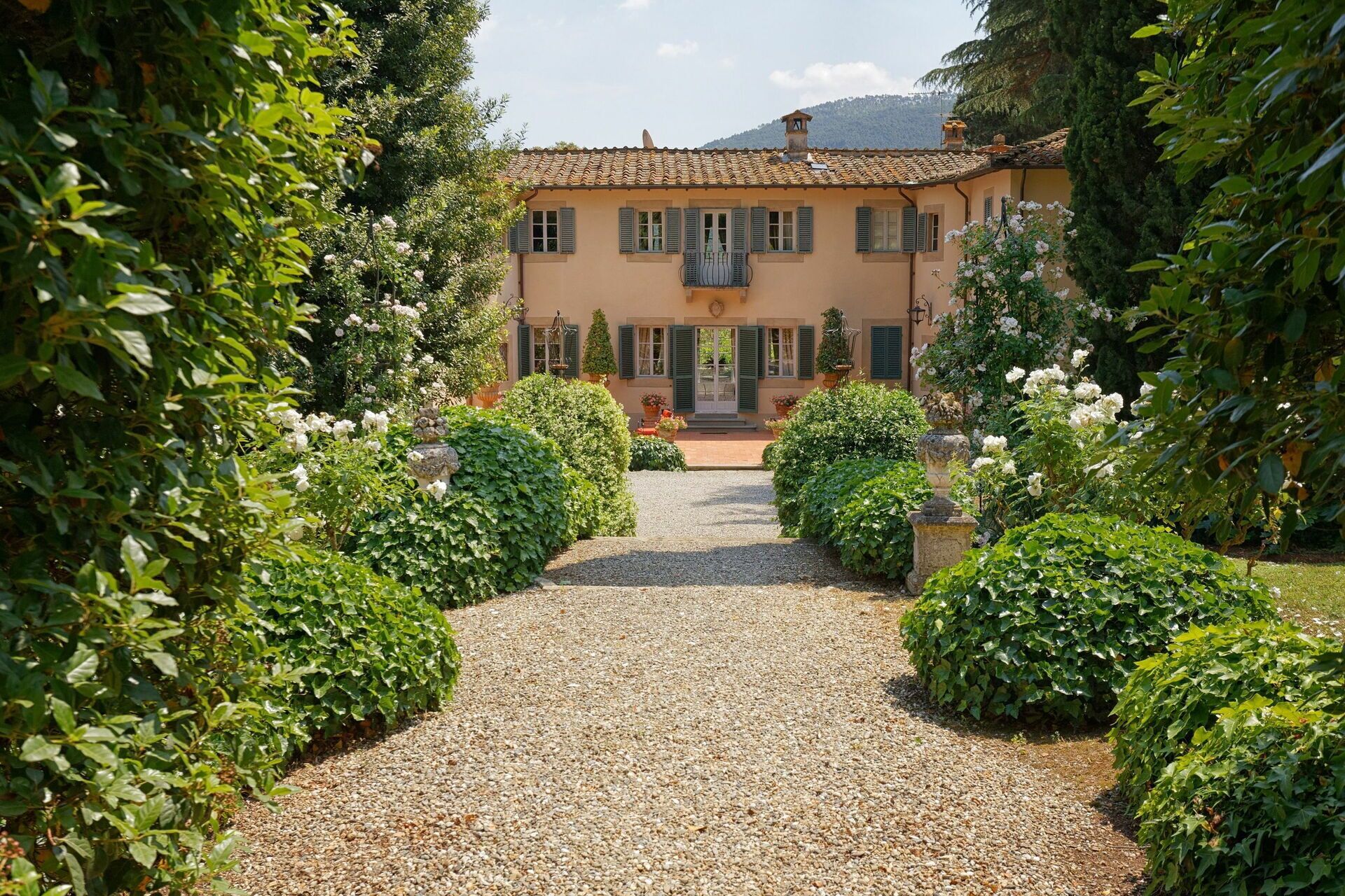 A villa with a gravel driveway leading to a front door, surrounded by lush greenery and white flowers.
