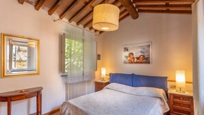 Una camera da letto con un letto, due abat-jour, un quadro, una finestra con tende leggere e un piccolo tavolo.