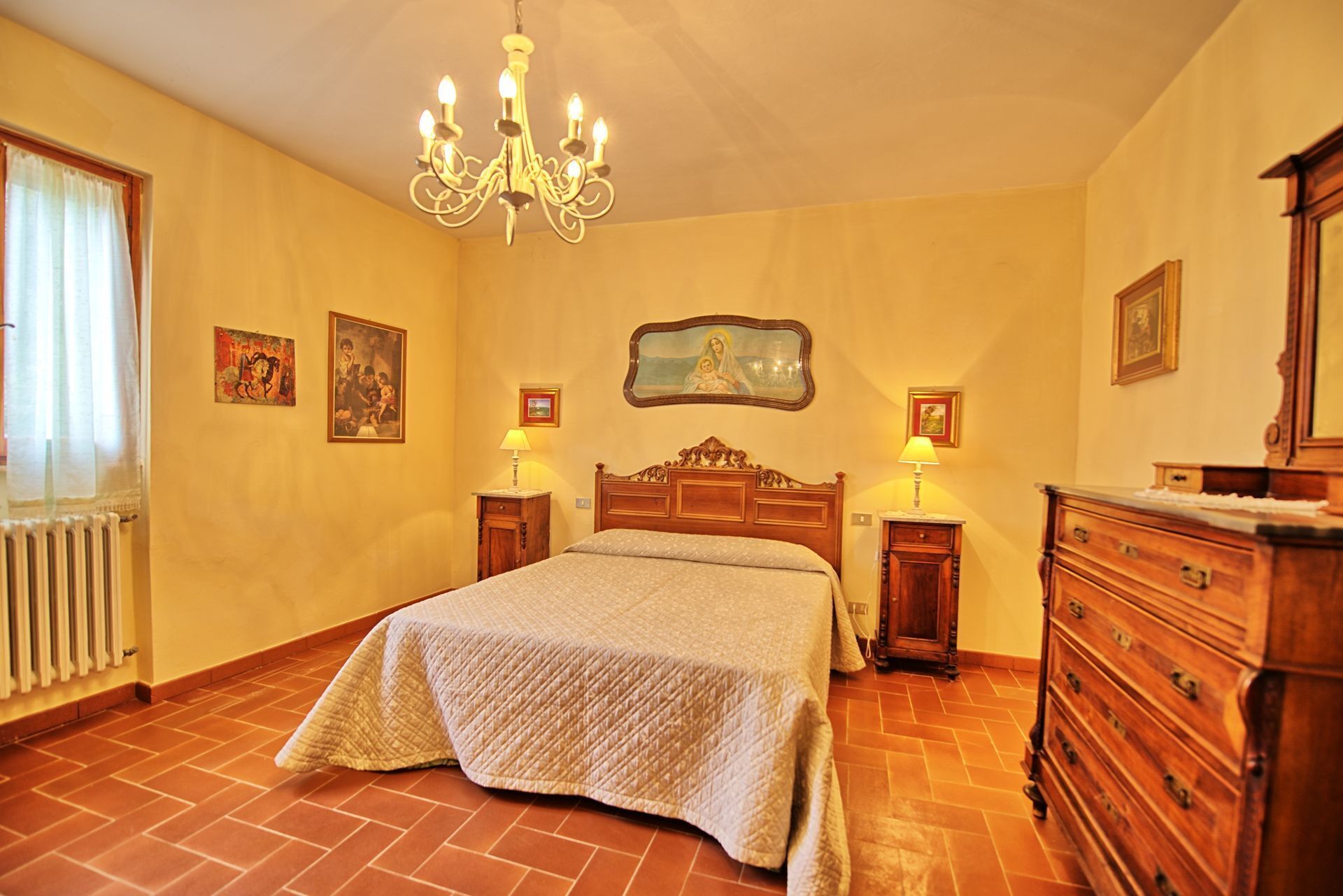 A bedroom with a large bed, wooden headboard, two bedside tables with lamps, a dresser, and a chandelier.