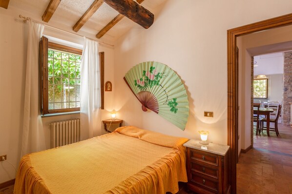 A bedroom with a bed, a bedside table with a lamp, a wall-mounted fan, and a window with curtains. - Cosy apartment in villa with WIFI, pool, TV, patio, pets allowed and panoramic view, close to Lu... (San Concordio Di Moriano)