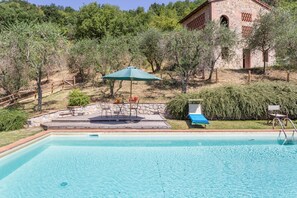 Ein Schwimmbecken mit Holzterrasse, Sitzgelegenheiten im Freien und einem Steinhaus im Hintergrund.