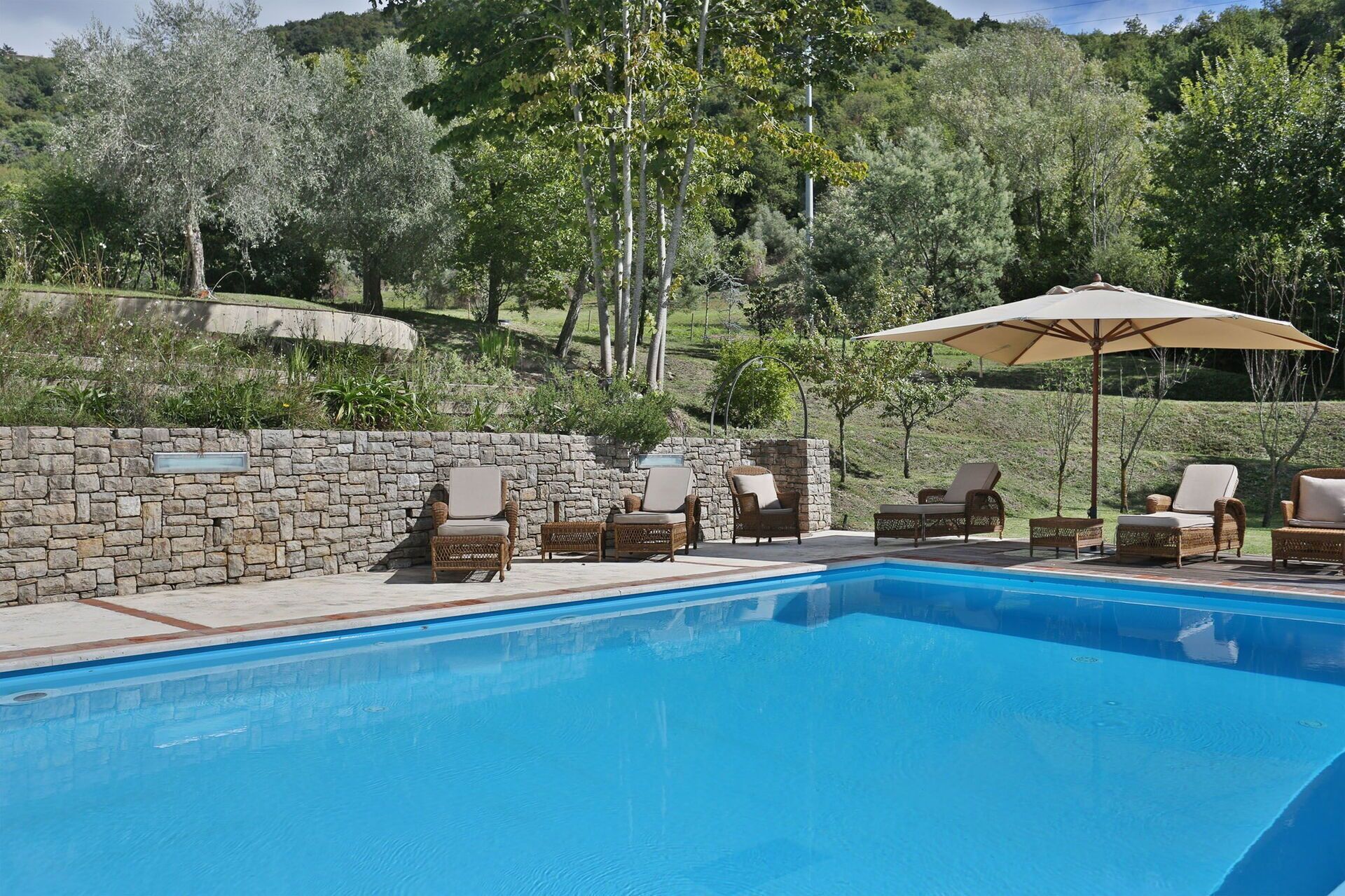 A swimming pool with lounge chairs and an umbrella, surrounded by a stone wall and greenery.