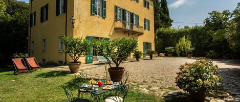 A yellow building with green shutters, a gravel courtyard, and a table with food and drinks.