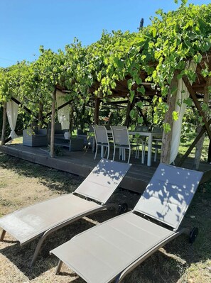 Uma pérgola com mesa e cadeiras sob ela, cercada por um jardim com um painel treliçado.