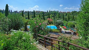 A lush garden with a swimming pool, a wooden deck, and a view of a hillside with buildings. - Beautiful apartment for 2 guests with pool, A/C, WIFI, TV and panoramic view (San Gimignano)