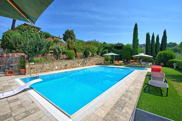 A rectangular swimming pool with a stone border, surrounded by a stone wall and a grassy area with outdoor seating.