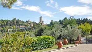 Un vialetto in pietra ciottolata fiancheggiato da vasi in terracotta e vegetazione rigogliosa, che conduce a una cittadina storica con edifici antichi e una torre imponente.