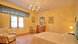 A bedroom with a bed, a chair, a dresser, a chandelier, and a window with curtains.