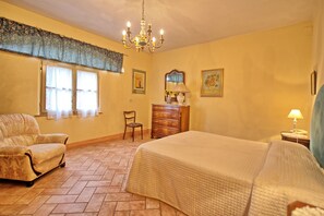 A bedroom with a bed, a chair, a dresser, a chandelier, and a window with curtains.