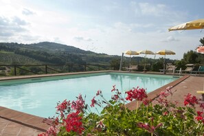 A swimming pool with lounge chairs and umbrellas, surrounded by a wooden deck and a view of green hills.