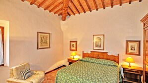 A bedroom with a wooden ceiling, a bed with a green patterned bedspread, a chair with a striped pillow, and two bedside tables with lamps. - Vittoriana 3 - Apartment with pool (San Gimignano)