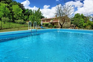 A swimming pool with clear blue water, surrounded by lush greenery and a brick building in the background.