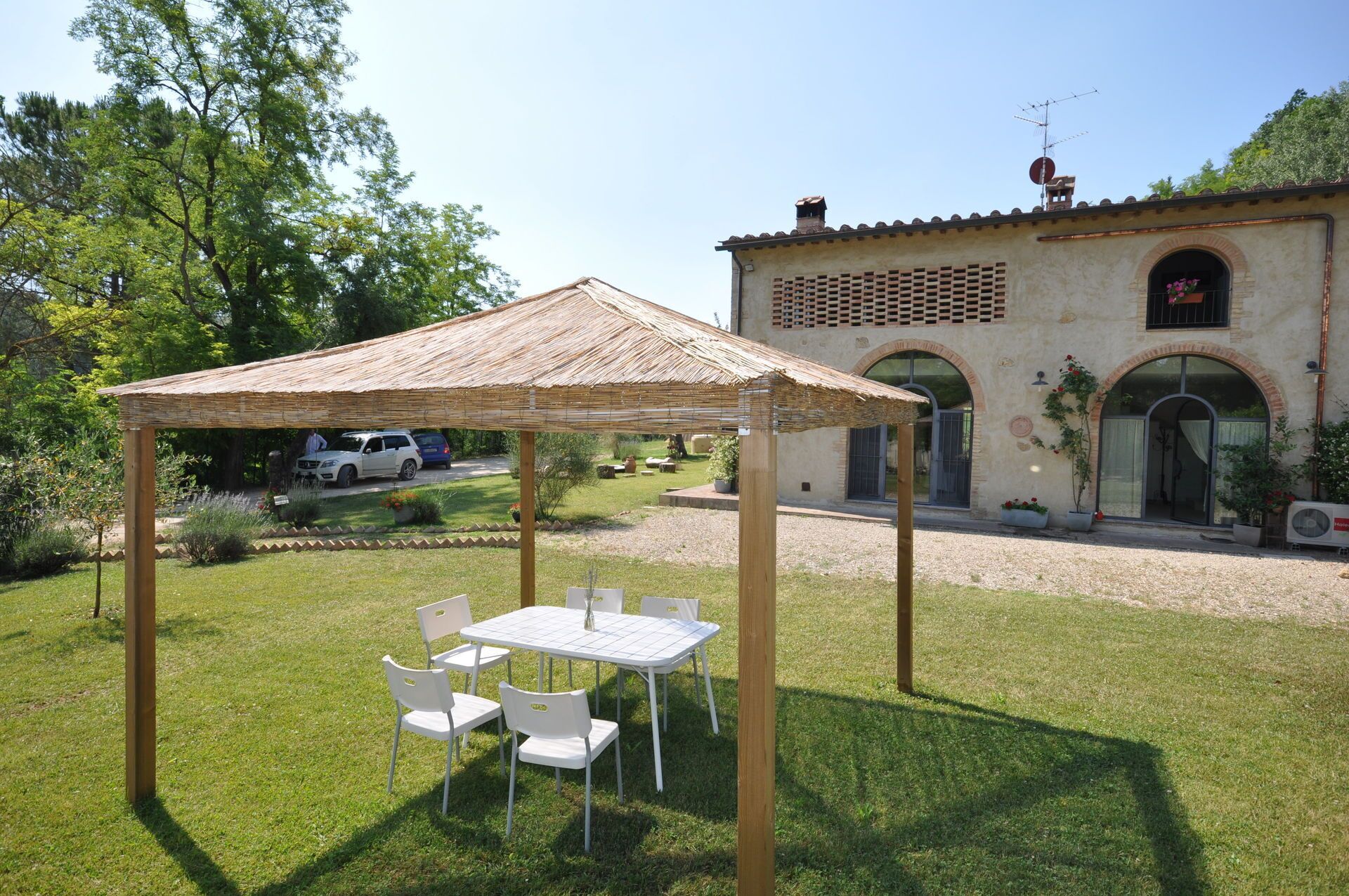 A gazebo with a table and chairs in a garden area, a building with arched windows and a chimney in the background.