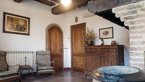 A rustic room with exposed wooden beams, a tiled floor, a wooden door, a cabinet with a vase of flowers, and a framed painting on the wall.