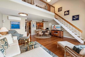 A spacious living area with a high ceiling, a dining table surrounded by chairs, a staircase, and a kitchen in the background.