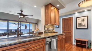 A modern kitchen with wooden cabinets, a stainless steel dishwasher, and a ceiling fan. - 🏖️Oceanfront Aliʻi Drive Condo w/ A/C | Walk to Town (Kailua Kona)