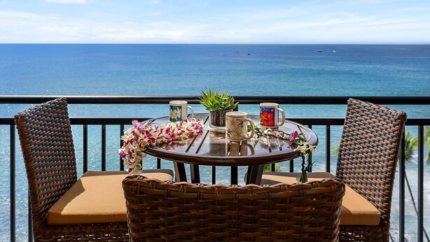 A round table with two wicker chairs, a potted plant, and two mugs on a balcony overlooking the ocean. - 🏖️Oceanfront Aliʻi Drive Condo w/ A/C | Walk to Town (Kailua Kona)