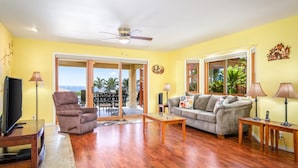 A living room with a sofa, armchair, and TV, wooden flooring, and a ceiling fan.