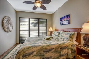 A bedroom with a bed, a ceiling fan, a window with blinds, a painting, and a decorative wall piece.