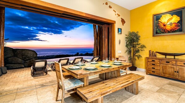 A dining area with a wooden table set for six, a large window with a sunset view, and a potted plant.