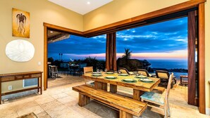 Un espace repas avec une grande table en bois, dressée pour six personnes, des bancs et une vue sur l’océan au coucher du soleil.
