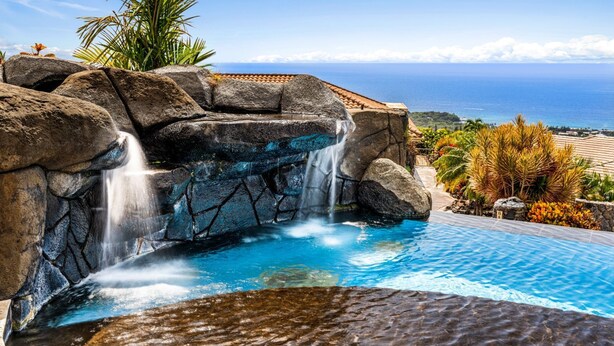 Une piscine agrémentée d’une cascade, entourée de plantes tropicales et offrant une vue sur l’océan.