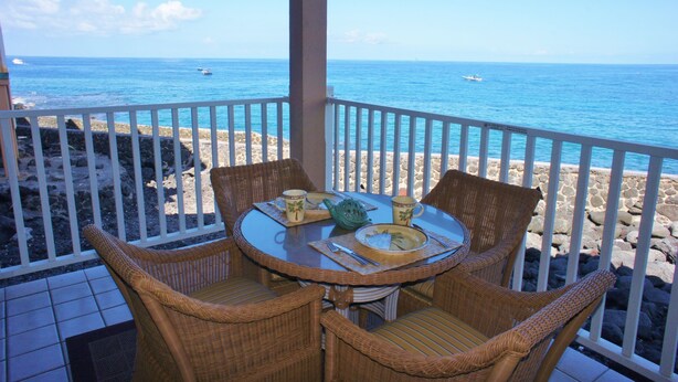 A covered outdoor dining area with wicker chairs and a table set for two, overlooking a rocky beach and the ocean. - 🏖️Oceanfront Kona Condo | Ground Floor, No Stairs (Kailua-Kona)