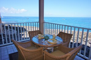 Un área de comedor al aire libre techada con sillas de mimbre y una mesa preparada para dos, con vista a una playa rocosa y el océano.