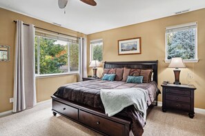 A bedroom with a large bed, two bedside tables, a ceiling fan, and a window with a view of trees.
