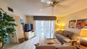 Ein Wohnzimmer mit einer Couch, einem Couchtisch und einem Fernseher. Eine Pflanze, eine Lampe und eine Uhr sind an der Wand zu sehen.
