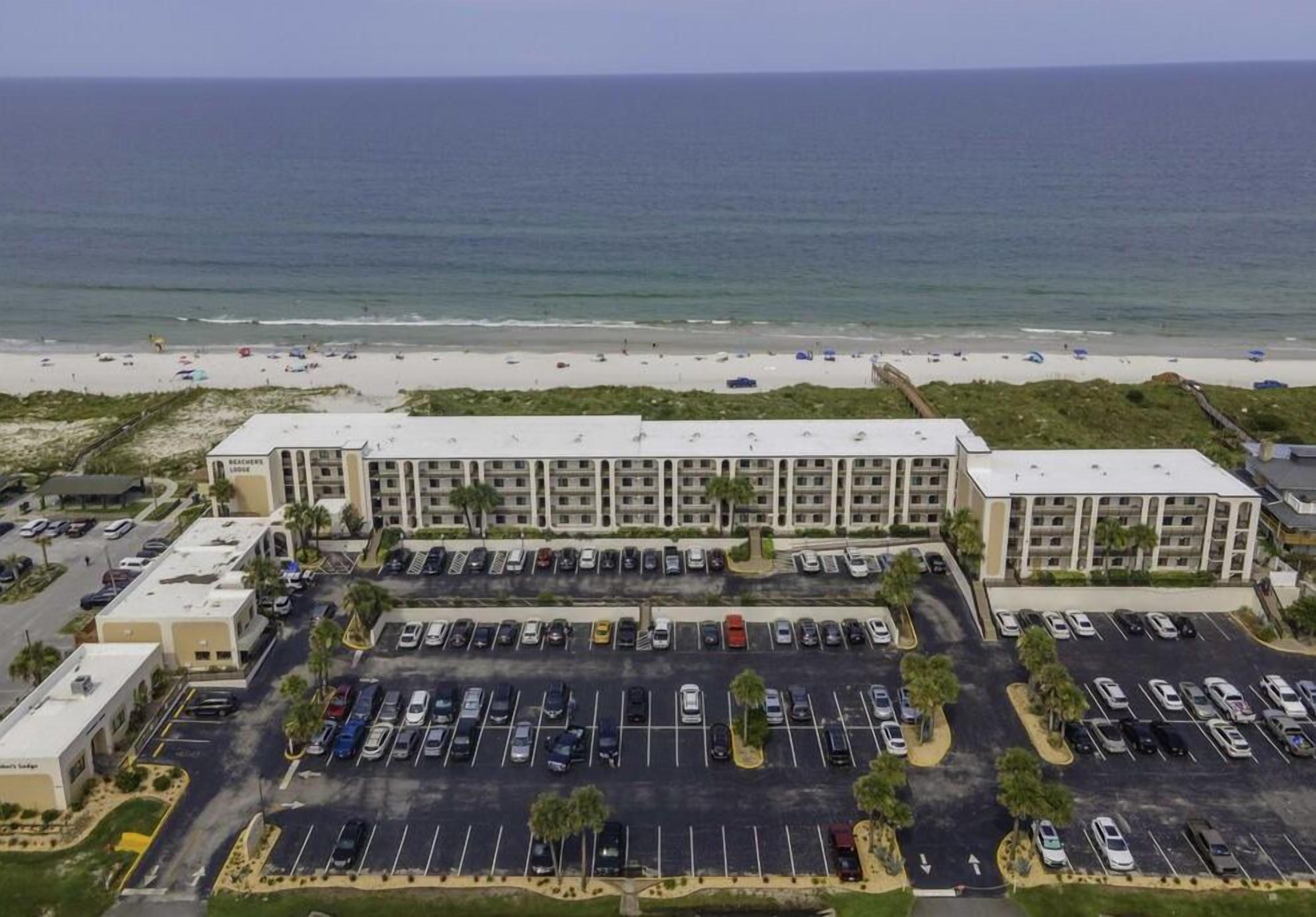 A multi-story hotel with a parking lot in front, overlooking a beach.