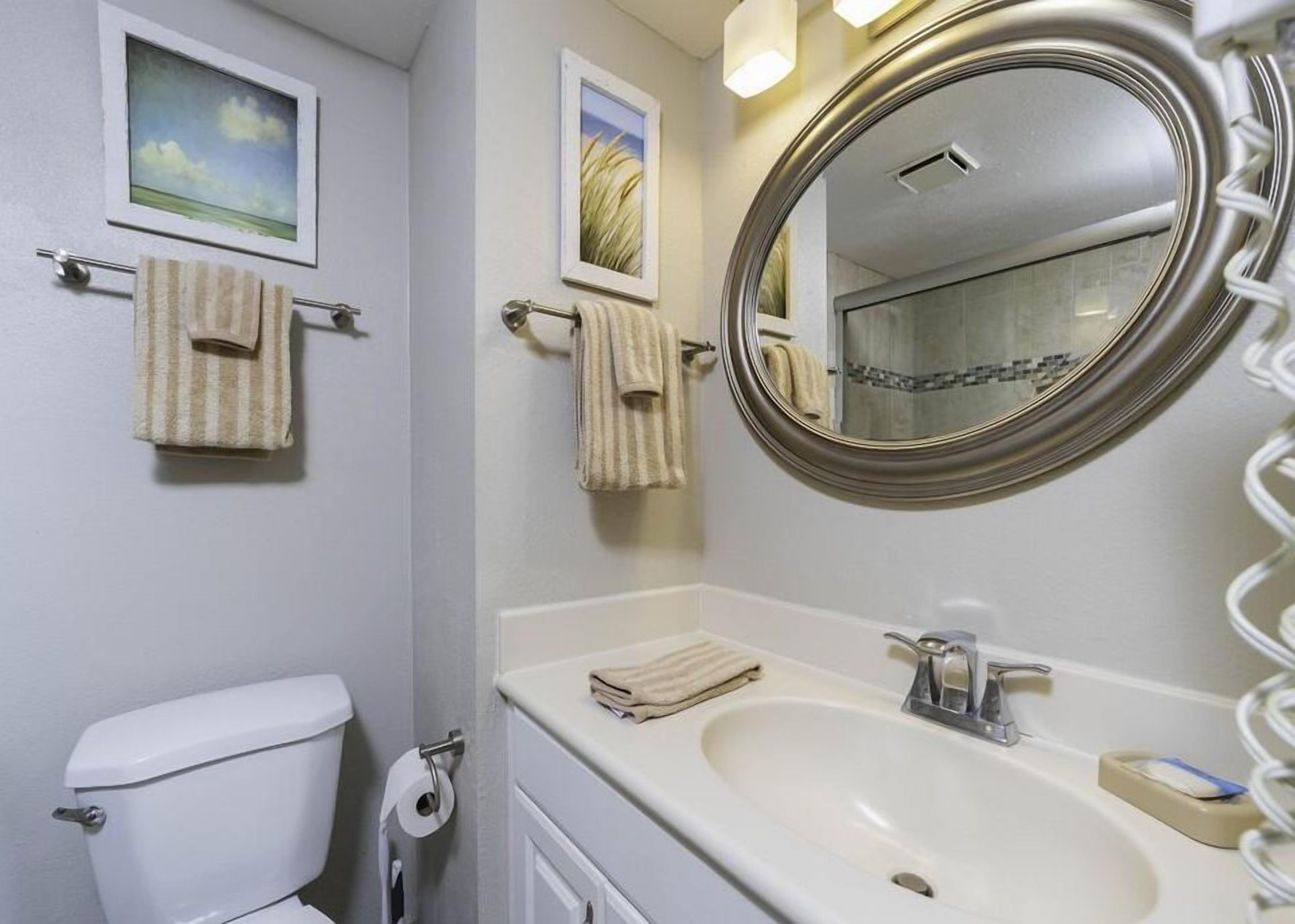 A bathroom with a toilet, sink, and mirror. There are towels hanging on towel bars and a picture on the wall.