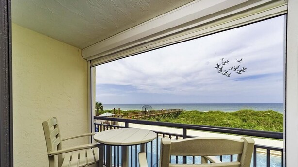 A balcony with two chairs and a small table, overlooking a beach with a wooden walkway and a clear sky. - Million Dollar Views ~ Heated Pool ~ King Bed (St Augustine)