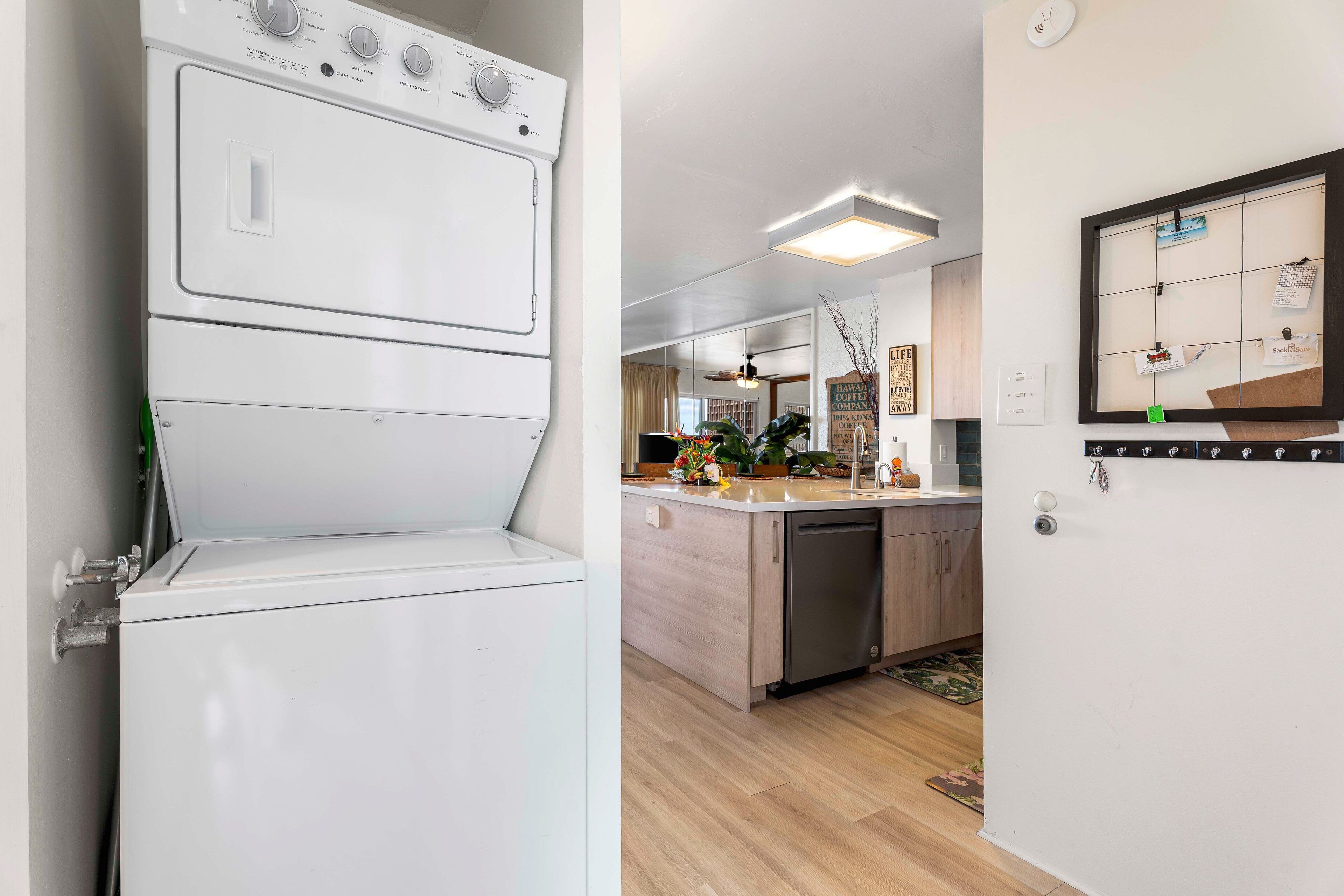 A modern kitchen with a dishwasher, a countertop with a sink, and a wall-mounted corkboard with notes.