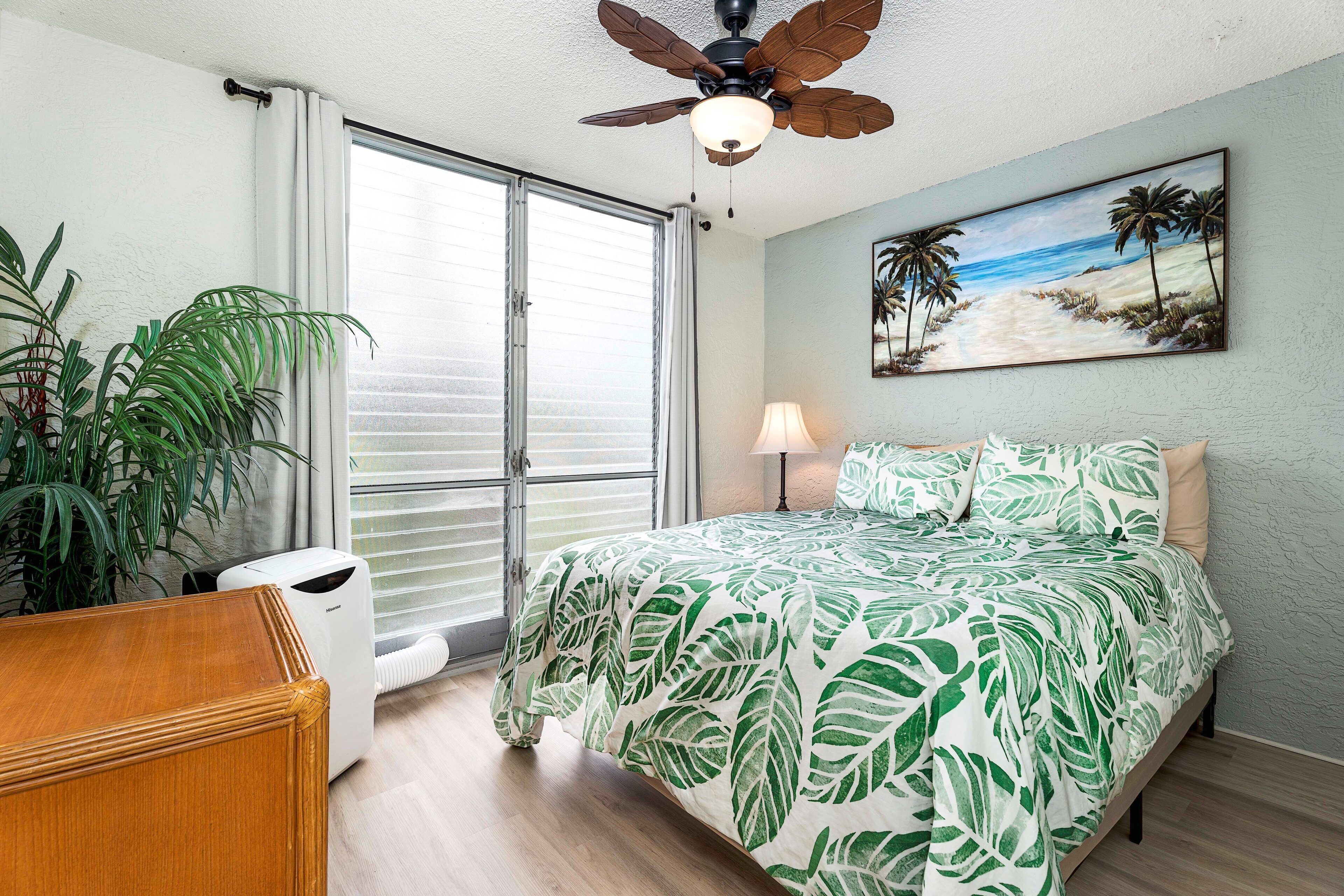 A bedroom with a bed, a ceiling fan, a painting of a beach, and a large window.