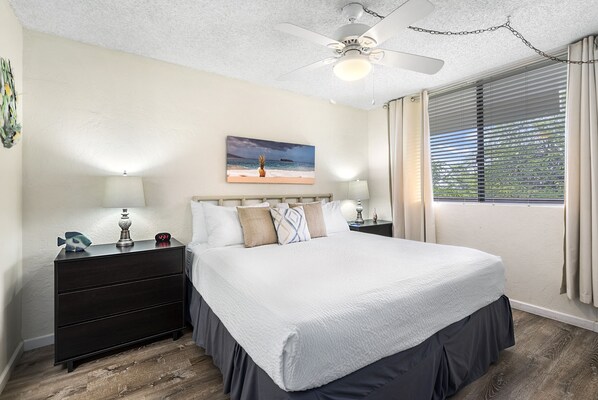 A bedroom with a bed, nightstands, a ceiling fan, a painting, and a window with blinds.