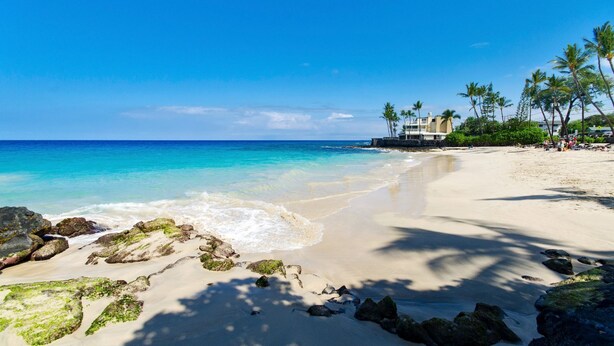 A beach with clear turquoise water, sandy shore, and a building with a balcony overlooking the sea.