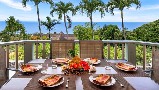 Mesa de comedor para seis personas, con platos, cubiertos y un centro de mesa con frutas, con vista a un paisaje marino con palmeras.
