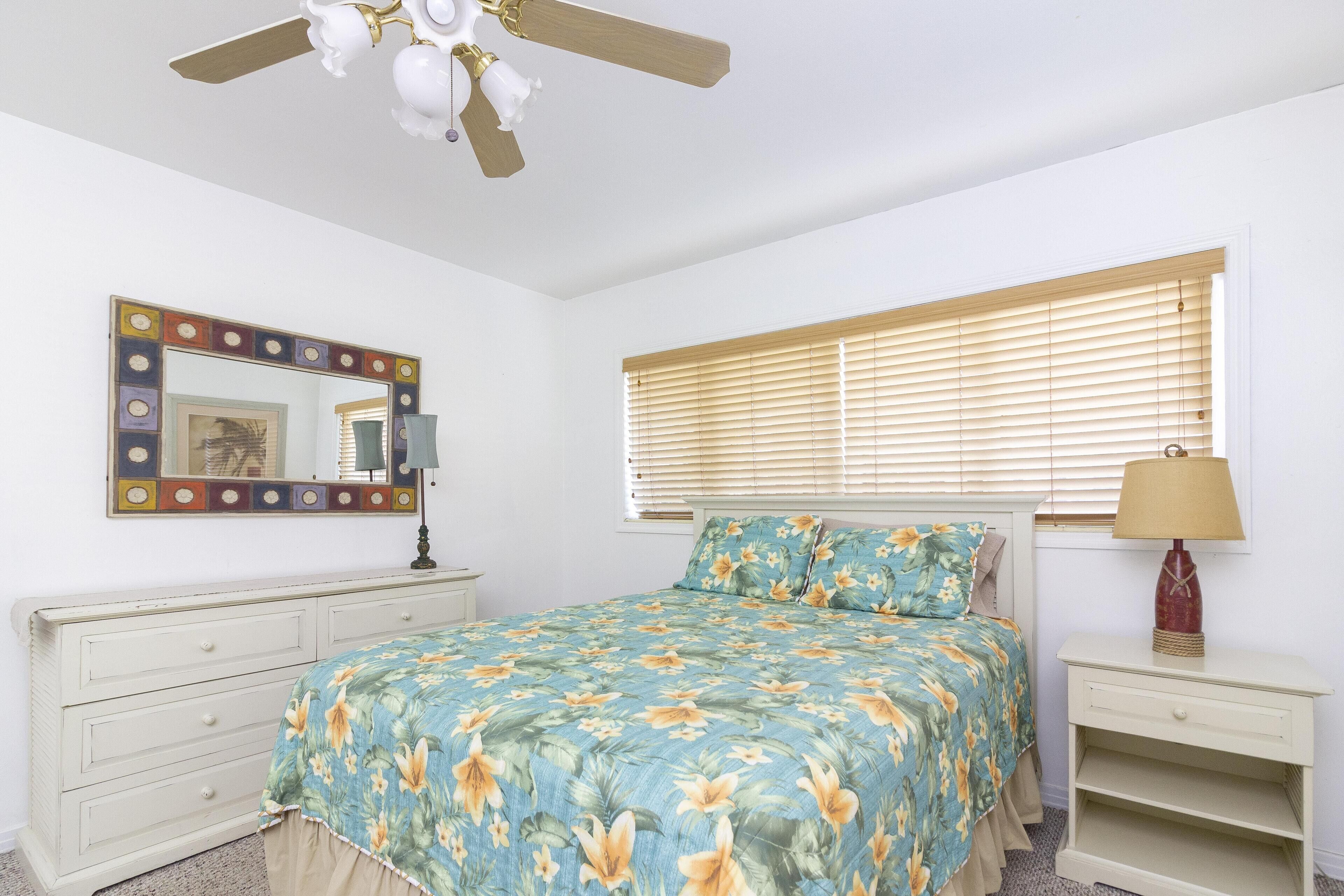 A bedroom with a bed, a dresser, a nightstand, a lamp, a ceiling fan, a window with blinds, and a mirror with a decorative frame.
