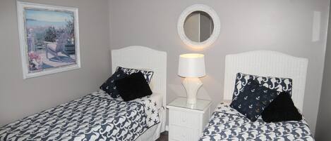 A bedroom with two beds, each with a patterned bedspread, a bedside lamp, a mirror, and a framed picture on the wall.