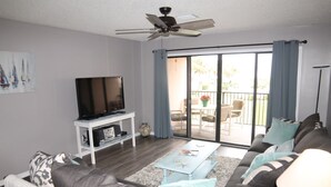 A living room with a sofa, coffee table, TV, and a balcony with outdoor furniture. - Sea Place 13239:  Charming Beach Condo: 2 Bedrooms, Pools & Tennis Courts (St. Augustine)