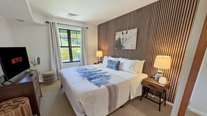 Un dormitorio ordenado con cama, lámparas de noche, un televisor mostrando 'NETFLIX', una ventana con cortinas y una obra de arte colgada en la pared.