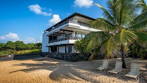 A two-story beachfront property with a balcony, a stone wall, and a palm tree.