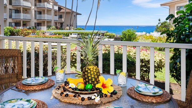 Et bord dækket med tallerkener, en ananas og tropisk frugt på en balkon med udsigt over en strand og hotelbygninger.