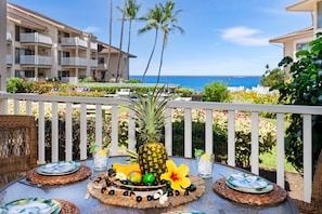A table set with plates, a pineapple, and tropical fruits on a balcony overlooking a beach and hotel buildings. - ποΈOcean View Ground Floor Kona Condo | Near Honl's (Kailua)