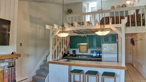 Une cuisine dotée d’un îlot central, de tabourets en bois, d’un escalier et d’un coin repas avec un plafond haut et des luminaires suspendus.